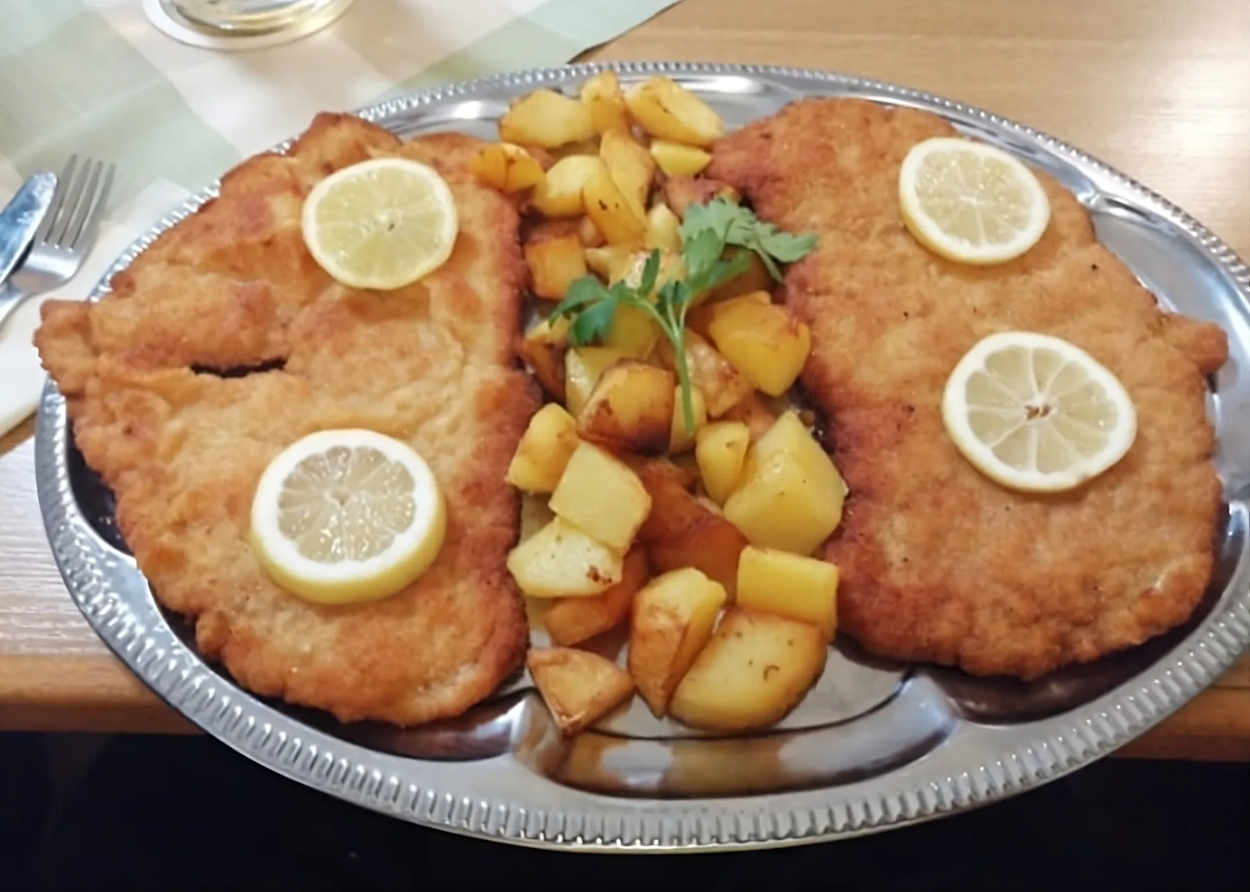 Two large breaded schnitzels with lemon slices and fried potatoes on a silver platter